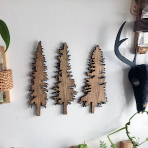 May include: Three wooden wall decor pieces shaped like pine trees. The trees are a light brown color and have a rustic, natural look.