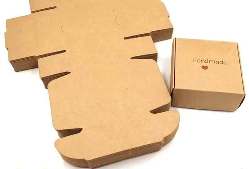 May include: A stack of brown cardboard boxes with a single box in the foreground. The box in the foreground has the word "Handmade" printed on it with a small red heart.
