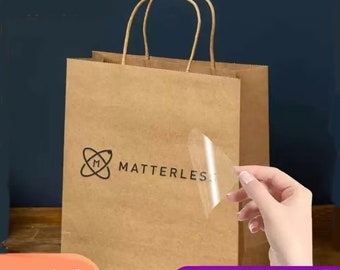 Può includere: Un sacchetto di carta marrone con un manico bianco e un adesivo trasparente con il testo "MATTERLESS" e un logo "M" stilizzato. L'adesivo viene staccato dal sacchetto.
