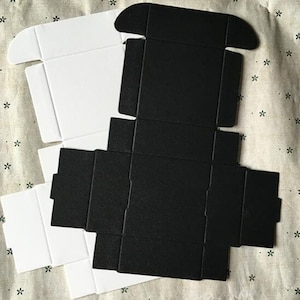 May include: Two flat pack boxes, one white and one black, are shown folded flat. The boxes are made of cardboard and have a simple design.