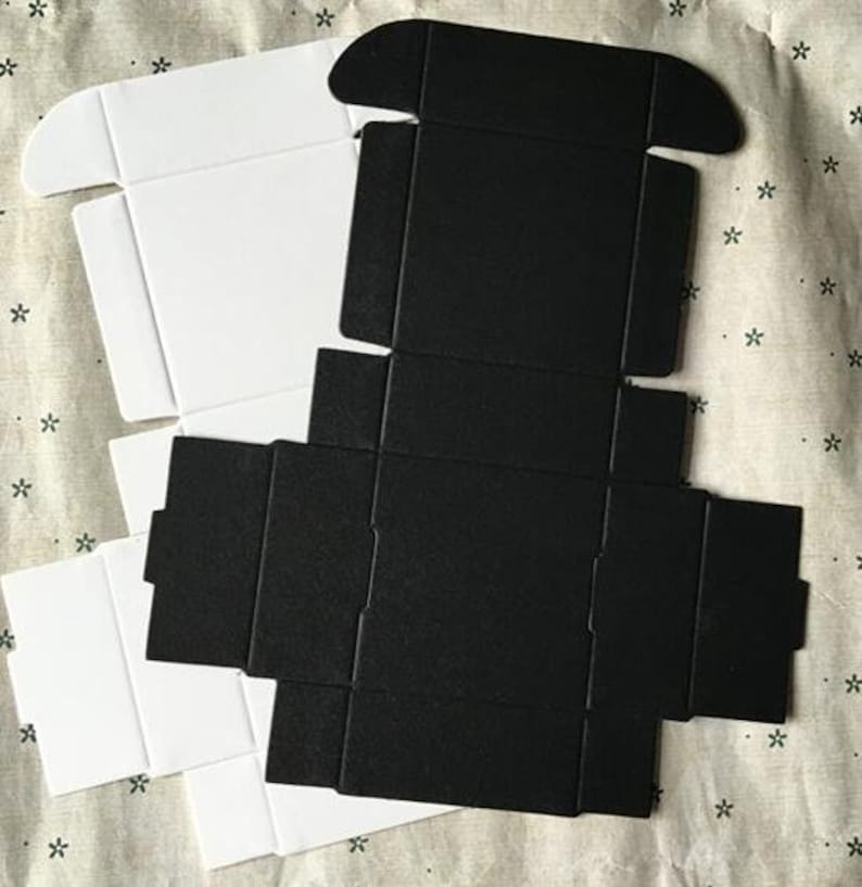 May include: Two flat pack boxes, one white and one black, are shown folded flat. The boxes are made of cardboard and have a simple design.