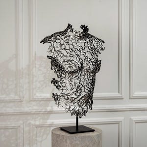 Puede incluir: Una escultura metálica de color plateado con forma de torso humano, construida a partir de una intrincada red calada de formas orgánicas interconectadas. La pieza se sostiene sobre una varilla vertical negra y una base cuadrada.