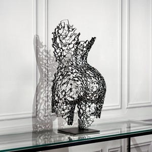 Könnte beinhalten: Eine Skulptur eines menschlichen Torsos aus einer dunklen, metallischen Gitterstruktur. Das Objekt besteht aus einem komplexen, miteinander verbundenen Netz aus geschwungenen Linien, die eine Silhouette bilden, und steht auf einem flachen, quadratischen Sockel aus schwarzem Metall.