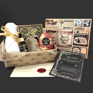 Könnte beinhalten: Ein Geschenkset in einer braunen Pappschachtel. Es enthält ein weißes Handtuch mit goldenem Band, eine schwarze Flasche, einen Jutesack, eine rot-gelbe Badekugel und ein Stück Seife. Eine dekorative Karte und ein Umschlag sind ebenfalls enthalten.