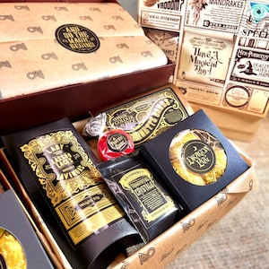 May include: A themed gift box containing various items such as Dragon Eggs, Lady Lasso Laces, and a red sweet. The box is adorned with vintage-style labels and the phrase "And So The Magic Begins."