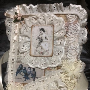 May include: A white lace journal featuring a vintage image of a woman in a white dress. The journal is adorned with a satin ribbon and lace and floral embellishments.