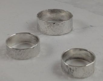 May include: Three silver rings with a hammered texture. The rings are wide band style and appear to be handmade. The rings are displayed on a white surface.