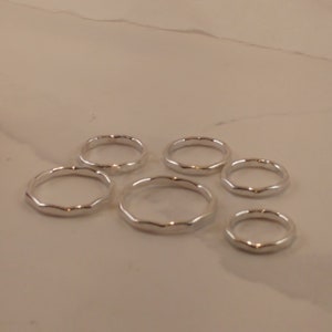 May include: Six silver rings with a wavy, textured design. The rings are displayed on a light-coloured surface. The rings are a simple, elegant design.