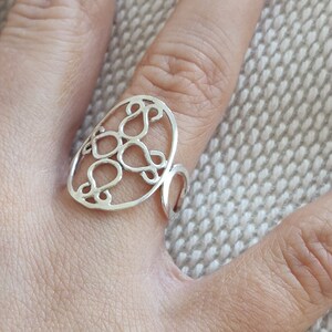 Peut inclure: Une bague en argent avec un motif filigrané ovale. La bague présente un motif ajouré de formes de goutte d'eau interconnectées à l'intérieur du cadre ovale. La bague est portée au doigt, mettant en valeur ses détails complexes et sa surface réfléchissante.