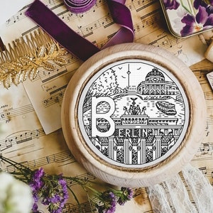 Op de afbeelding: Een ronde houten ornament met een zwart-wit Berlijns stadsgezicht ontwerp, met de Brandenburger Tor en de letter "B". Het ornament is omgeven door vintage items zoals bladmuziek, bloemen en een gouden haarkam.