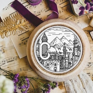 Könnte beinhalten: Eine Schwarzweißillustration der Skyline von Kairo mit dem Wort "Cairo" darunter. Die Illustration befindet sich auf einem runden Holzsockel und ist von Notenblättern, Blumen und anderen Dekorationsartikeln umgeben.