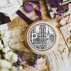 Op de afbeelding: Een witte houten borduurring met een zwart-wit ontwerp van de Eiffeltoren en de Notre Dame kathedraal in Parijs, Frankrijk. De ring is omgeven door vintage muziekbladen, kant en paars lint.