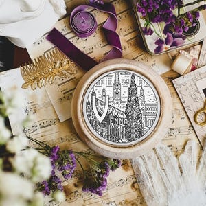 May include: A round, wooden framed art piece with a black and white illustration of a city skyline, with the word "ENNA" in the center. The artwork is surrounded by vintage items, including sheet music, flowers, and gold scissors.