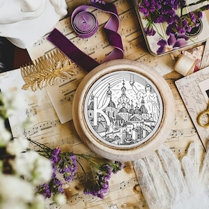 Może przedstawiać: Białe drewniane tamborek do haftu z czarno-białym wzorem miejskiego krajobrazu i napisem "KYIV" w środku. Tamborek leży na stole z nutami, kwiatami i innymi ozdobami.