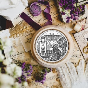 May include: A white wooden round embroidery hoop with a black and white design of a city skyline and the word "MADRID" in a banner. The hoop is on a table with music sheets, flowers, and other decorative items.