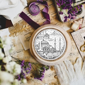 Op de afbeelding: Een ronde houten onderzetter met een zwart-witte illustratie van de skyline van Istanbul, inclusief de Hagia Sophia en de Galata-toren. De onderzetter is omringd door vintage bladmuziek, kant en bloemen.