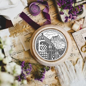 Op de afbeelding: Een witte houten borduurring met een zwart-witte lijntekening van een gebouw met de tekst "EDINBURGH" eronder. De ring is omgeven door muziekbladen, bloemen en andere decoratieve items.
