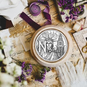 Op de afbeelding: Een witte borduurring met een zwart-witte illustratie van het Vrijheidsbeeld en het Empire State Building. De tekst "NEW YORK" staat onder het Vrijheidsbeeld. De ring ligt op een tafel met bladmuziek, bloemen en andere knutselspullen.