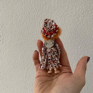 Puede incluir: Una peque&ntilde;a mu&ntilde;eca con pelo naranja que lleva un vestido blanco con manchas marrones y naranjas y un sombrero rojo y blanco. La mu&ntilde;eca lleva un broche plateado en la parte posterior de su vestido.