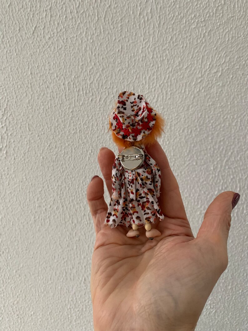 Puede incluir: Una peque&ntilde;a mu&ntilde;eca con pelo naranja que lleva un vestido blanco con manchas marrones y naranjas y un sombrero rojo y blanco. La mu&ntilde;eca lleva un broche plateado en la parte posterior de su vestido.