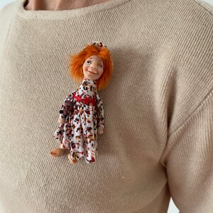 Puede incluir: Una peque&ntilde;a mu&ntilde;eca hecha a mano con cabello naranja, vestida con un vestido floral rojo y blanco y un sombrero rojo. La mu&ntilde;eca sonr&iacute;e y tiene una expresi&oacute;n alegre.