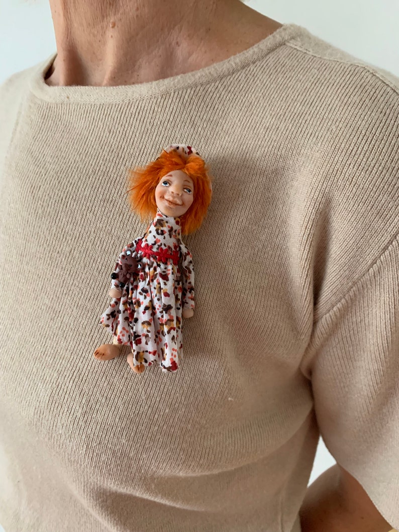 Puede incluir: Una peque&ntilde;a mu&ntilde;eca hecha a mano con cabello naranja, vestida con un vestido floral rojo y blanco y un sombrero rojo. La mu&ntilde;eca sonr&iacute;e y tiene una expresi&oacute;n alegre.