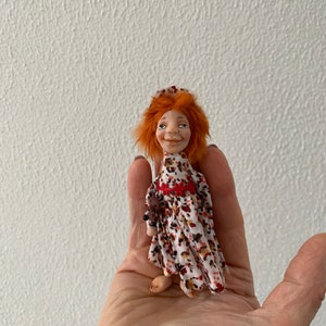 Puede incluir: Una mu&ntilde;eca hecha a mano con cabello rojo, vestida con un vestido blanco y rojo con estampado floral. La mu&ntilde;eca sostiene un peque&ntilde;o animal marr&oacute;n en su mano.