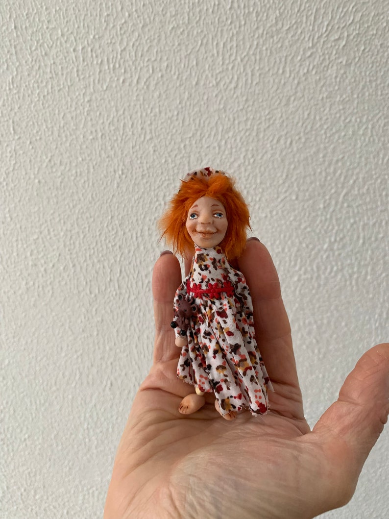 Puede incluir: Una mu&ntilde;eca hecha a mano con cabello rojo, vestida con un vestido blanco y rojo con estampado floral. La mu&ntilde;eca sostiene un peque&ntilde;o animal marr&oacute;n en su mano.