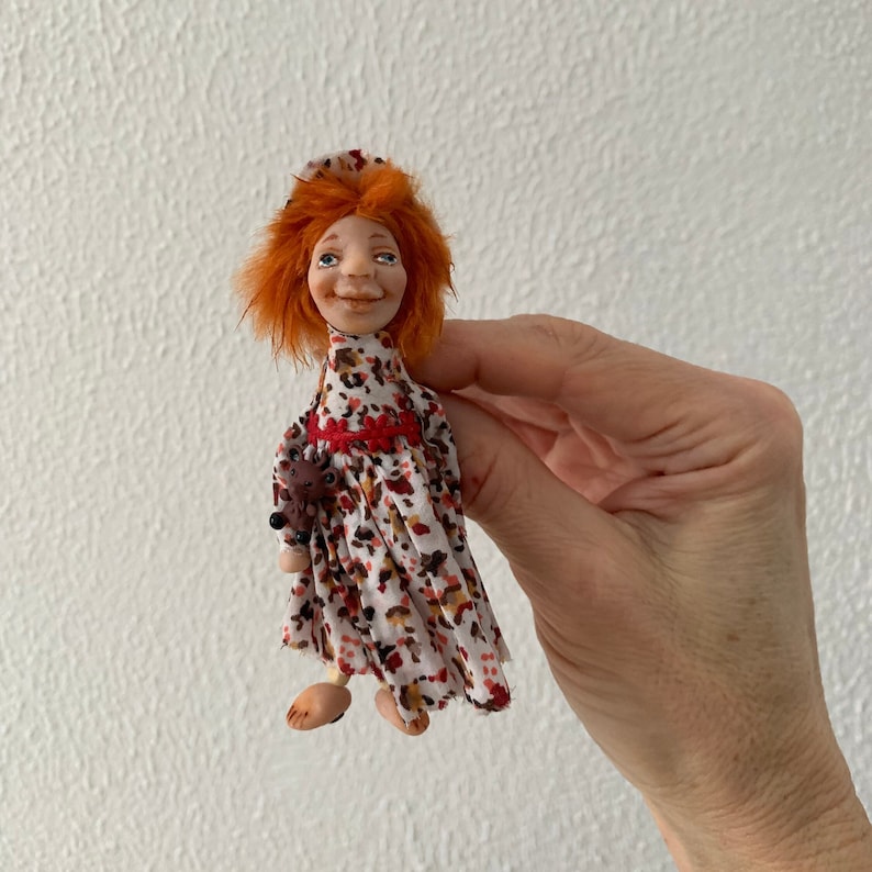 Puede incluir: Una mu&ntilde;eca hecha a mano con pelo rojo, vestida con un vestido estampado en blanco y marr&oacute;n con una cinta roja. La mu&ntilde;eca lleva un peque&ntilde;o animal marr&oacute;n en sus brazos.