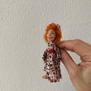 Puede incluir: Una peque&ntilde;a mu&ntilde;eca con cabello rojo que lleva un vestido blanco con estampado floral marr&oacute;n y rojo. La mu&ntilde;eca sostiene un peque&ntilde;o oso de peluche marr&oacute;n.