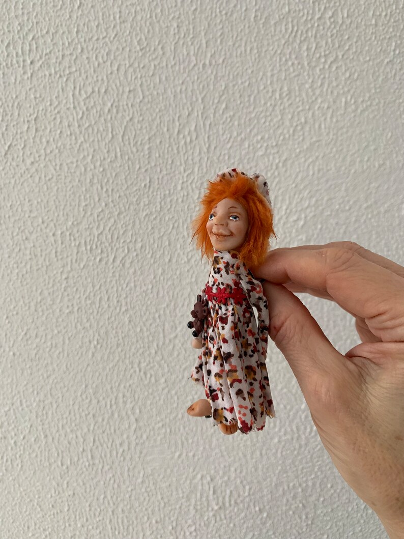 Puede incluir: Una peque&ntilde;a mu&ntilde;eca con cabello rojo que lleva un vestido blanco con estampado floral marr&oacute;n y rojo. La mu&ntilde;eca sostiene un peque&ntilde;o oso de peluche marr&oacute;n.