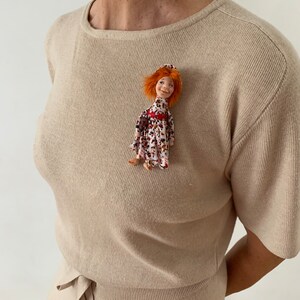 Puede incluir: Un su&eacute;ter de punto beige con un peque&ntilde;o broche de mu&ntilde;eca. La mu&ntilde;eca tiene el pelo rojo, un sombrero rojo y un vestido floral.