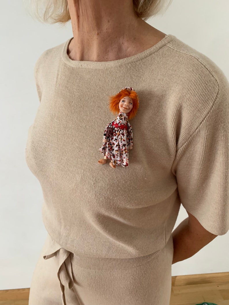 Puede incluir: Un su&eacute;ter de punto beige con un peque&ntilde;o broche de mu&ntilde;eca. La mu&ntilde;eca tiene el pelo rojo, un sombrero rojo y un vestido floral.