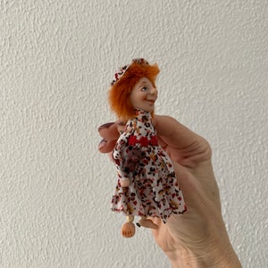 Puede incluir: Una mu&ntilde;eca peque&ntilde;a con pelo rojo, vestida con un vestido blanco con estampado floral rojo y marr&oacute;n, un sombrero rojo y piernas de madera.