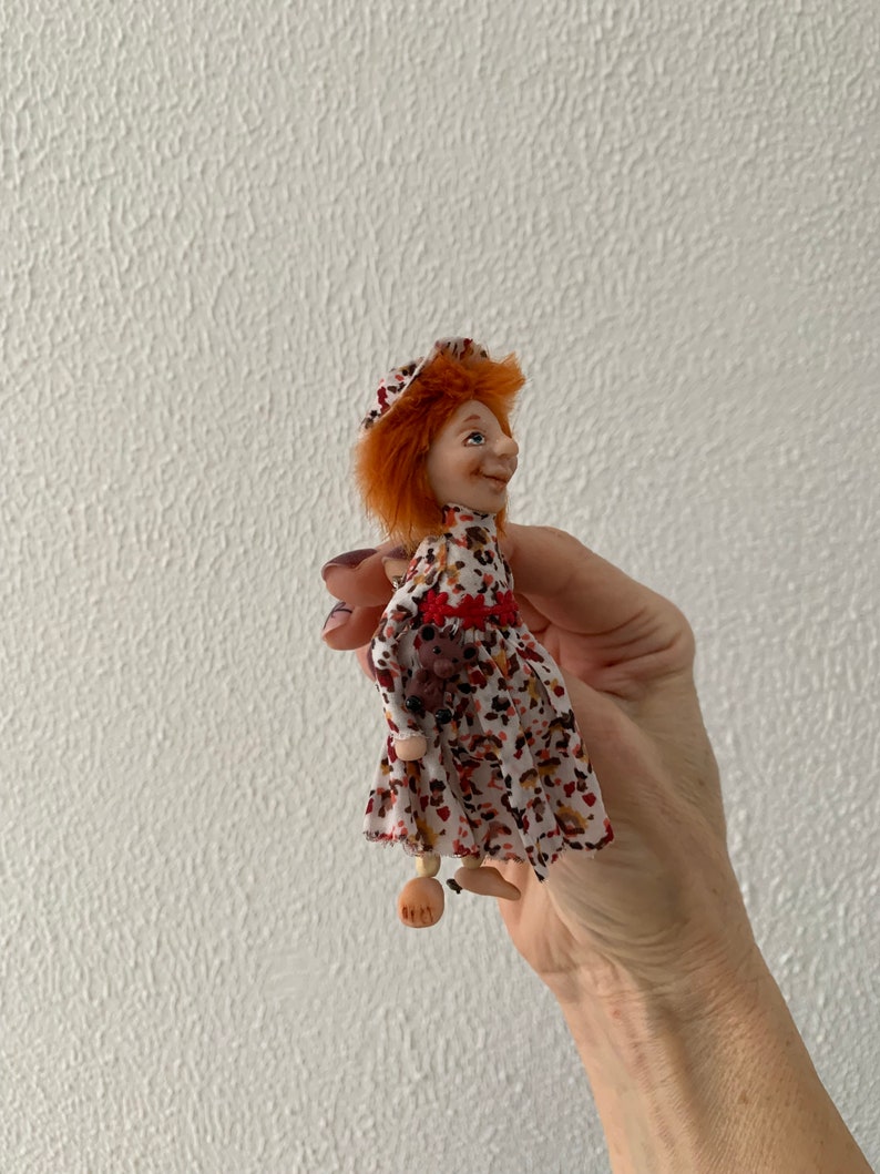 Puede incluir: Una mu&ntilde;eca peque&ntilde;a con pelo rojo, vestida con un vestido blanco con estampado floral rojo y marr&oacute;n, un sombrero rojo y piernas de madera.