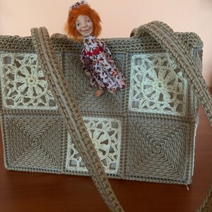 Puede incluir: Un bolso de crochet con un patr&oacute;n marr&oacute;n y blanco. El bolso tiene una correa larga y est&aacute; hecho de cuadrados. Una peque&ntilde;a mu&ntilde;eca con cabello rojo est&aacute; sentada encima del bolso.