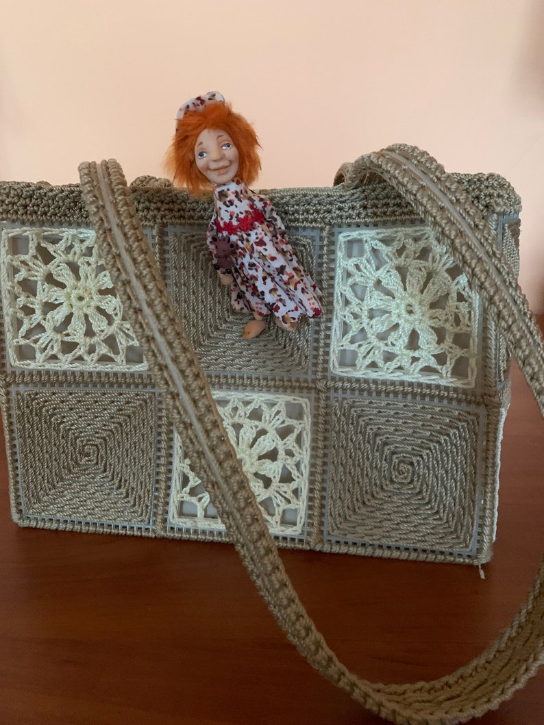 Puede incluir: Un bolso de crochet con un patr&oacute;n marr&oacute;n y blanco. El bolso tiene una correa larga y est&aacute; hecho de cuadrados. Una peque&ntilde;a mu&ntilde;eca con cabello rojo est&aacute; sentada encima del bolso.