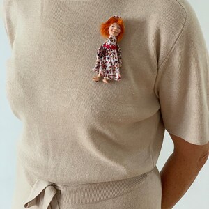 Puede incluir: Una blusa de punto beige de manga corta con un lazo en la cintura. Una peque&ntilde;a mu&ntilde;eca con el pelo rojo y un vestido floral est&aacute; prendida en la parte delantera de la blusa.