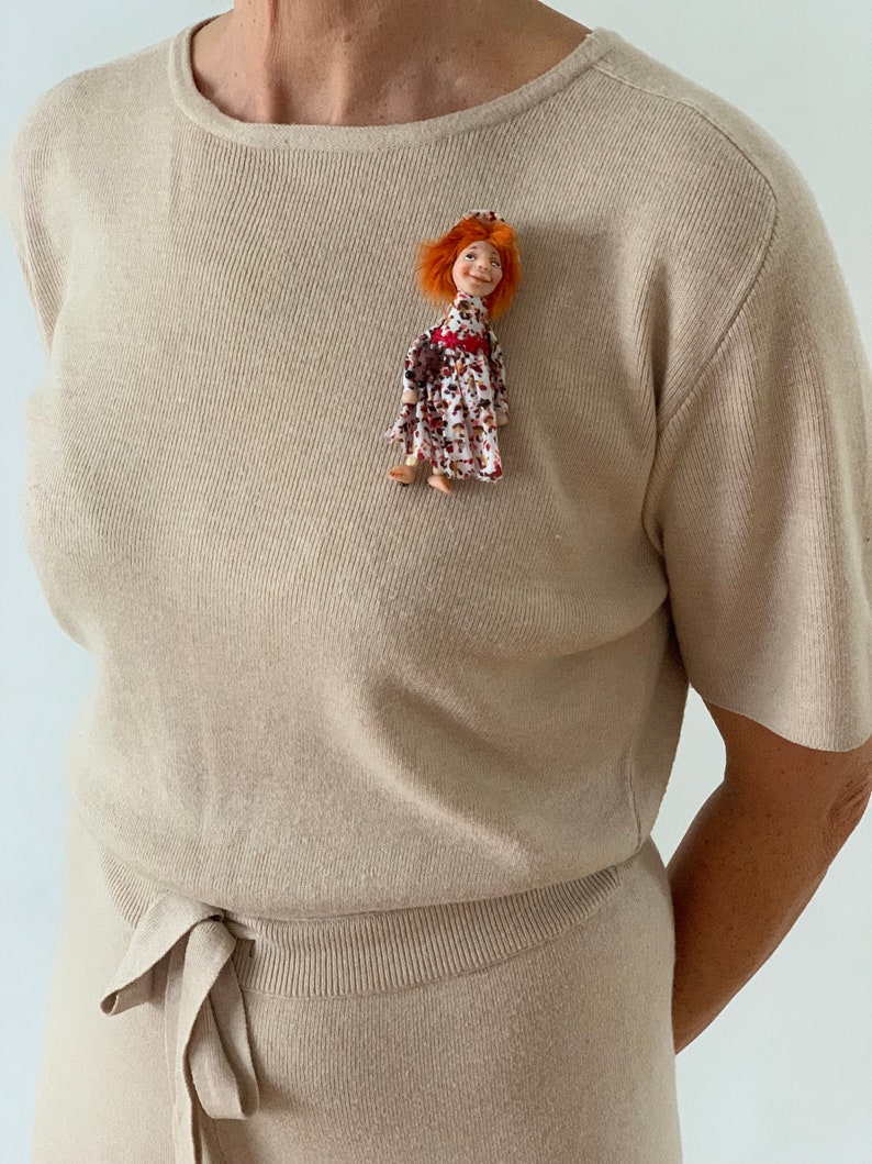 Puede incluir: Una blusa de punto beige de manga corta con un lazo en la cintura. Una peque&ntilde;a mu&ntilde;eca con el pelo rojo y un vestido floral est&aacute; prendida en la parte delantera de la blusa.