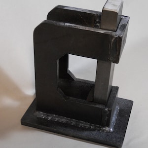 May include: A black metal press with a square opening and a metal bar that slides up and down. The press is mounted on a black metal base.