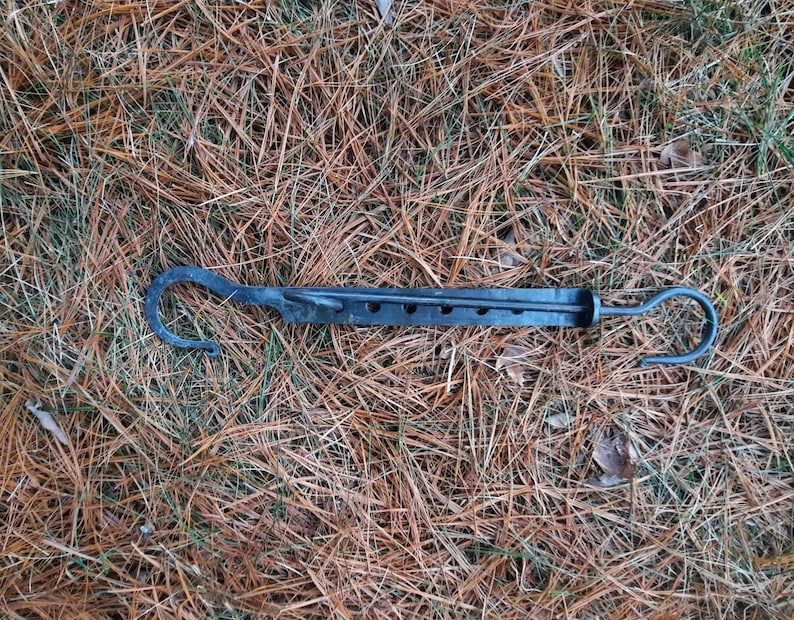 Peut inclure: Un outil de chemin&eacute;e en m&eacute;tal noir avec un crochet &agrave; chaque extr&eacute;mit&eacute; et une s&eacute;rie de trous sur toute sa longueur. L'outil est pos&eacute; sur un lit d'aiguilles de pin brunes.