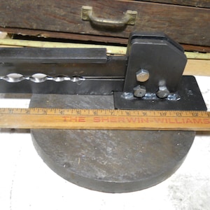 May include: A black metal tool with a handle and a series of holes for shaping metal. The tool is mounted on a round metal base. A wooden ruler with the text "THE SHERWIN-WILLIAMS CO." is placed in front of the tool.