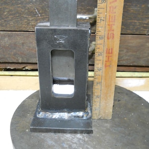 May include: A black metal tool with a rectangular opening and a sliding metal piece. The tool is sitting on a round, gray surface. The tool is 6 inches tall.