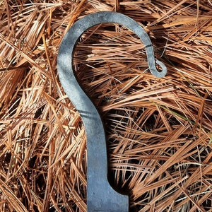 Peut inclure: Un crochet en m&eacute;tal au design unique. Le crochet est en m&eacute;tal fonc&eacute; et a une forme incurv&eacute;e lisse. Le crochet est pos&eacute; sur un lit d'aiguilles de pin brunes.