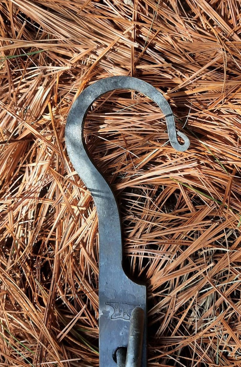 Peut inclure: Un crochet en m&eacute;tal au design unique. Le crochet est en m&eacute;tal fonc&eacute; et a une forme incurv&eacute;e lisse. Le crochet est pos&eacute; sur un lit d'aiguilles de pin brunes.