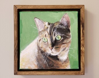 Puede incluir: Una pintura enmarcada de un gato con ojos verdes. El gato es marrón, negro y blanco y mira directamente al espectador. El fondo es de un color verde sólido.