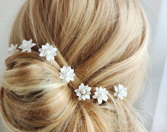 May include: Six white flower hair clips with pearl accents. The clips are arranged in a row in a woman's blonde hair.