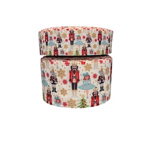 May include: Two rolls of white ribbon with a repeating Christmas-themed pattern. The pattern includes nutcrackers, ballerinas, snowflakes, and Christmas trees in red, blue, and gold. The ribbon is likely used for gift wrapping or crafting.