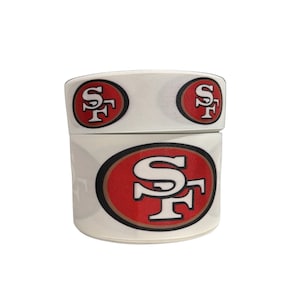 May include: A roll of white ribbon featuring the San Francisco 49ers logo. The logo is a red oval with a black outline, containing the letters "SF" in white. The ribbon is suitable for crafting and sports-themed projects.