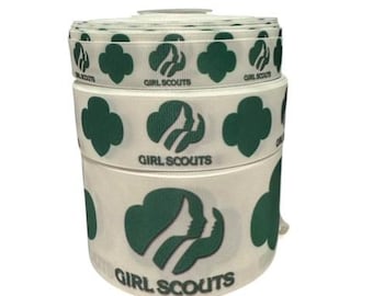 May include: A stack of white ribbon rolls featuring the Girl Scouts logo in dark green. The logo includes a profile of a girl and a trefoil. The words "GIRL SCOUTS" are printed on the ribbon.